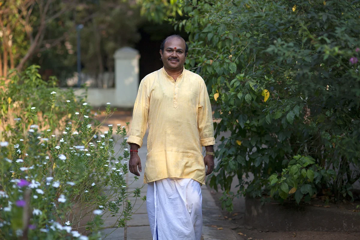 A man wearing traditional Indian attire, including a yellow kurta and white dhoti, walking along a garden path surrounded by greenery and flowers.