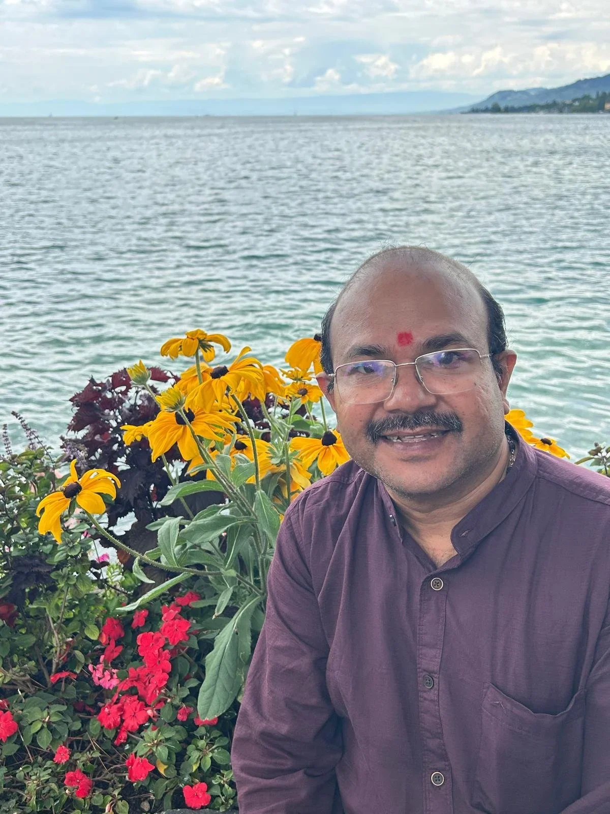 A man with glasses and a red mark on his forehead smiling outdoors near a body of water with yellow and pink flowers in the background.