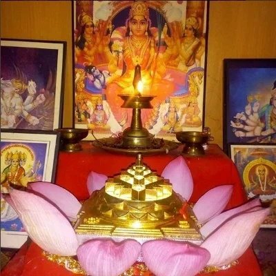 Hindu altar with a brass lamp, pink lotus flowers, and a small golden sculpture, surrounded by framed pictures of Hindu deities in the background.