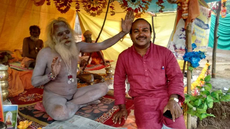 A man in red traditional clothing sitting on the ground with a bald head, smiling, in a tent decorated with flowers, and a dusty ground outside. An elderly man with long white beard and hair, wearing jewelry, is sitting cross-legged, raising one hand