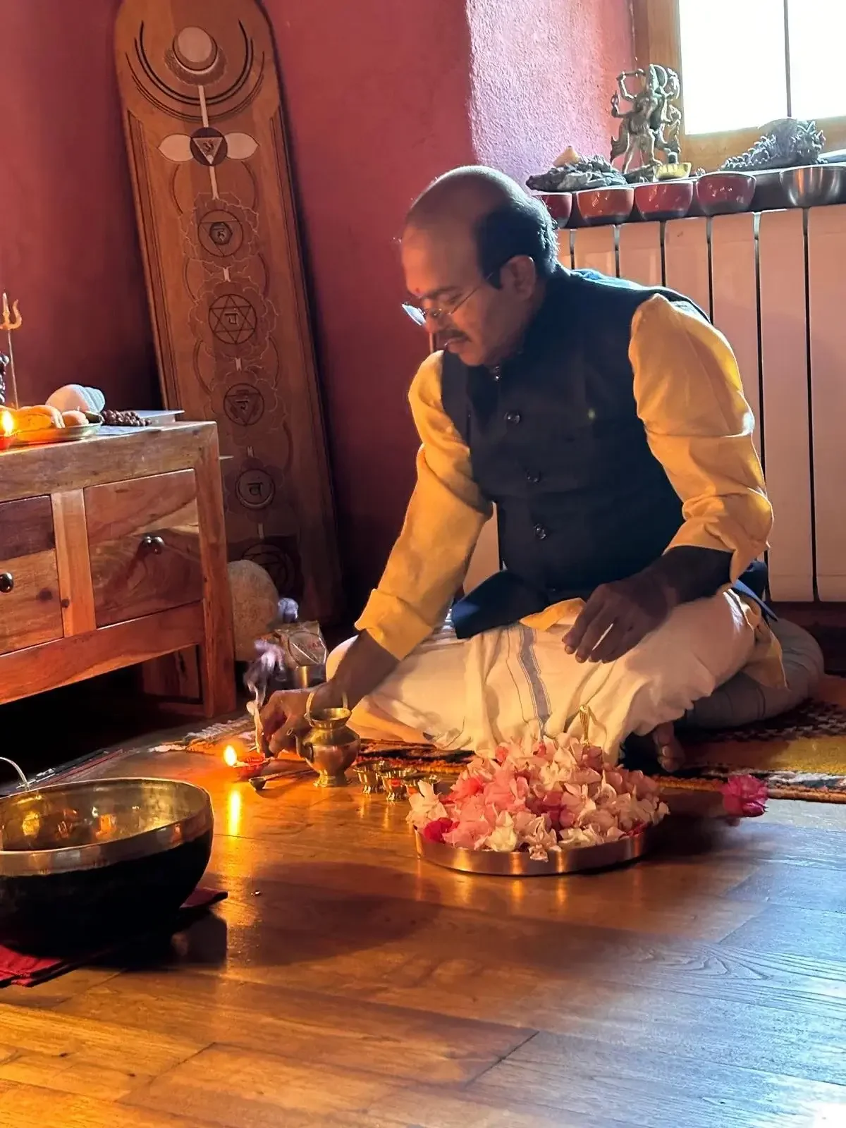 A man dressed in traditional Indian attire, kneeling on a rug, participating in a ritual or ceremony in a room with wooden furniture and decorative items. He is handling a brass vessel with pink flower petals around it, and a lit oil lamp is nearby.