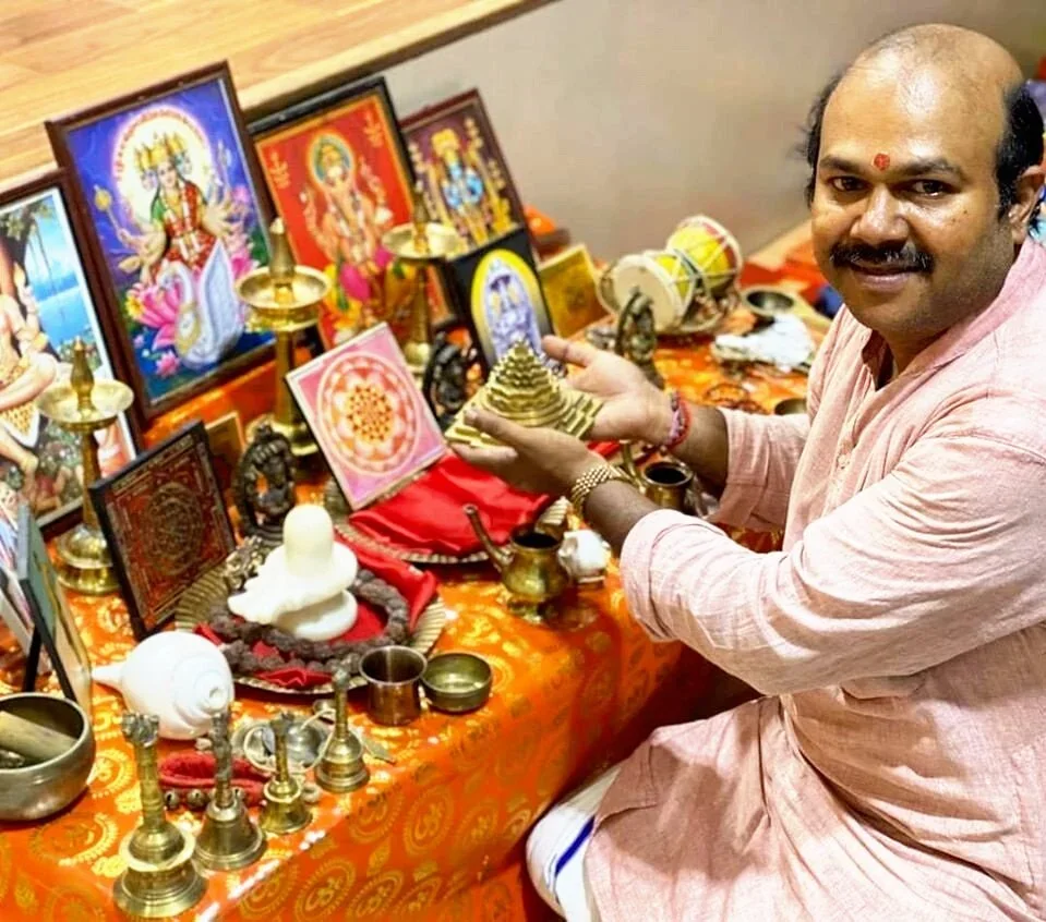 A man participating in a Hindu ritual, sitting at an altar with various religious images, statues, and offerings.