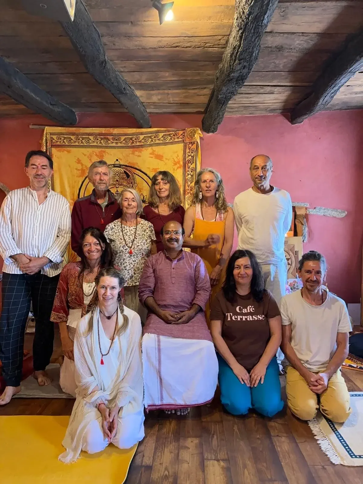 A group of fifteen people, some seated and some standing, gathered in a cozy room with wooden beams and pink walls. They are smiling and dressed casually, with some wearing traditional or spiritual attire. The background features a decorative yellow 
