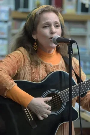 Young woman with light brown hair wearing an orange top and patterned dress, singing into a microphone while playing an acoustic guitar in a bookstore or library setting.