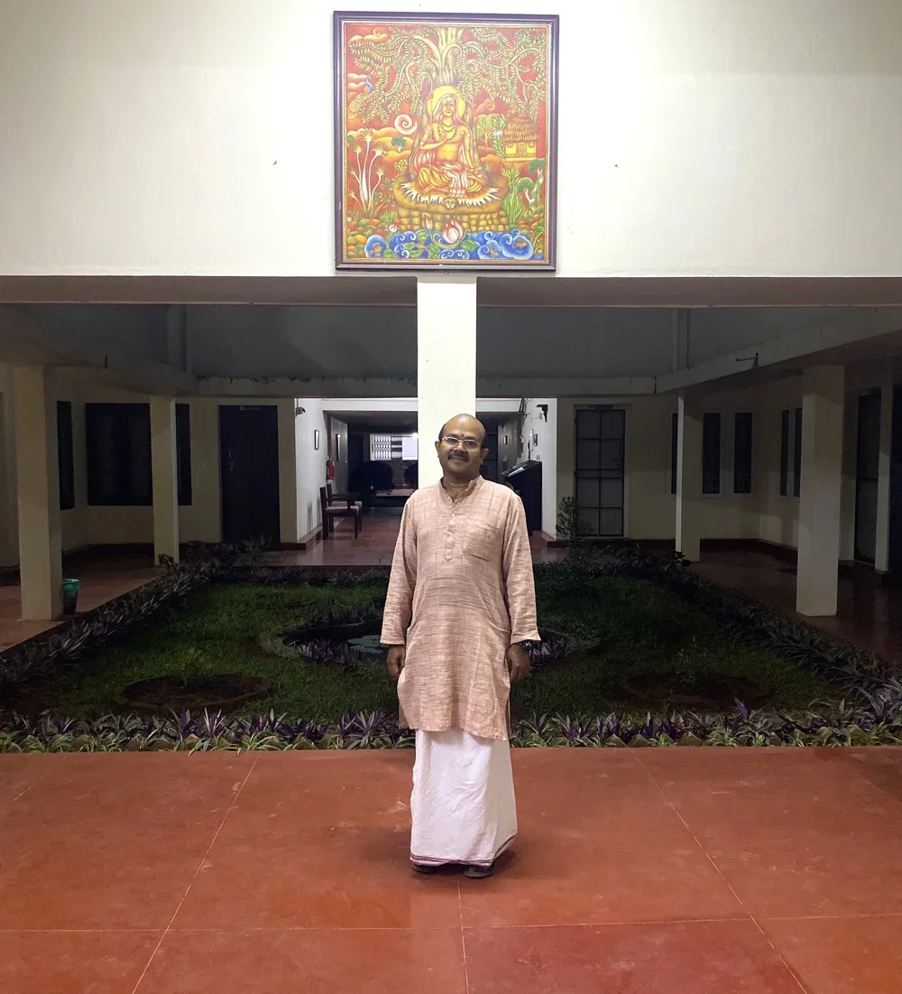 A man standing in a corridor with a garden behind him, in front of a white pillar. A colorful framed artwork depicting a deity is hung above the pillar.