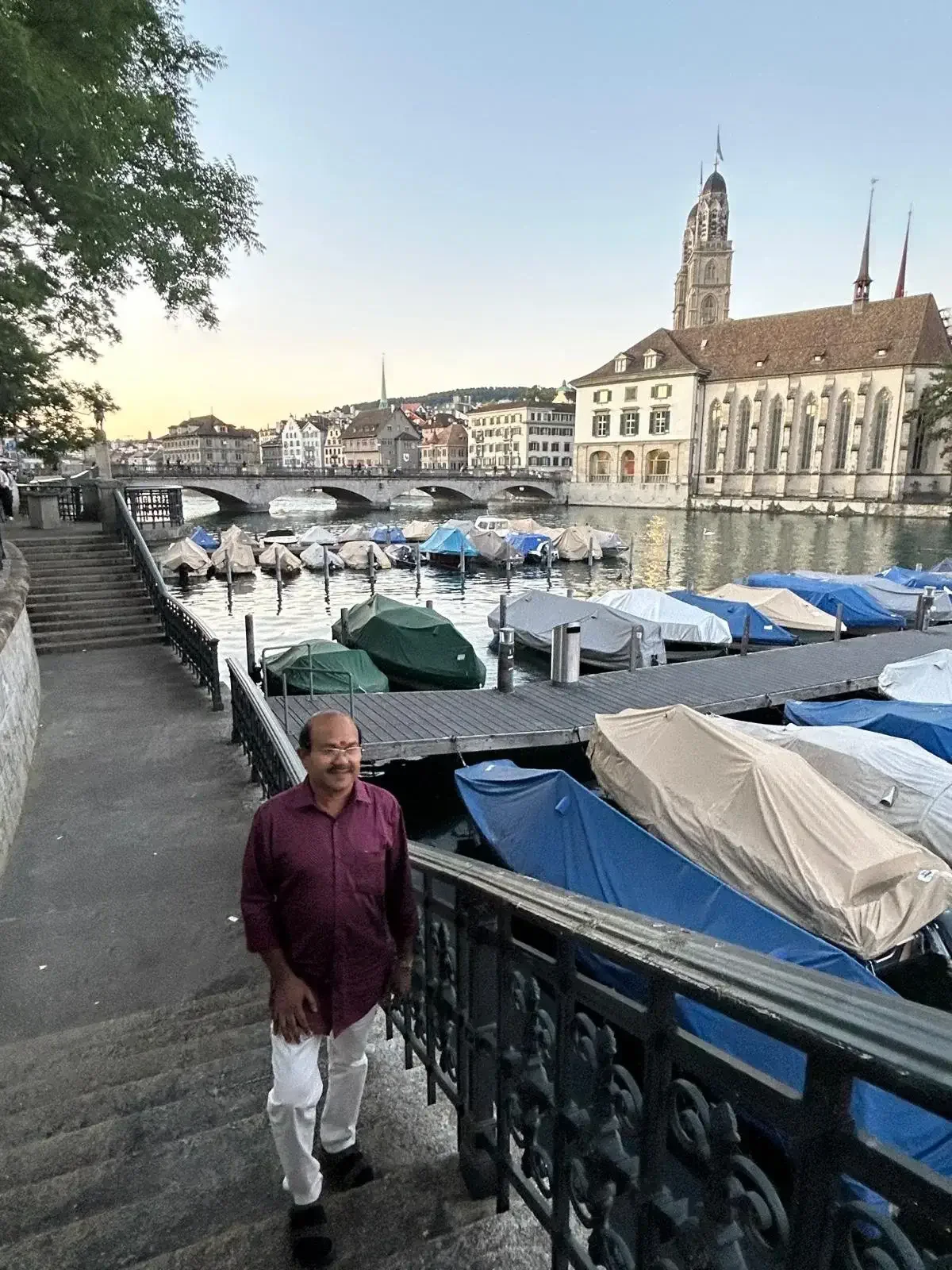 A man standing on stairs beside a river with covered boats docked along the edge, historic European buildings, and a large church with a tall spire in the background.
