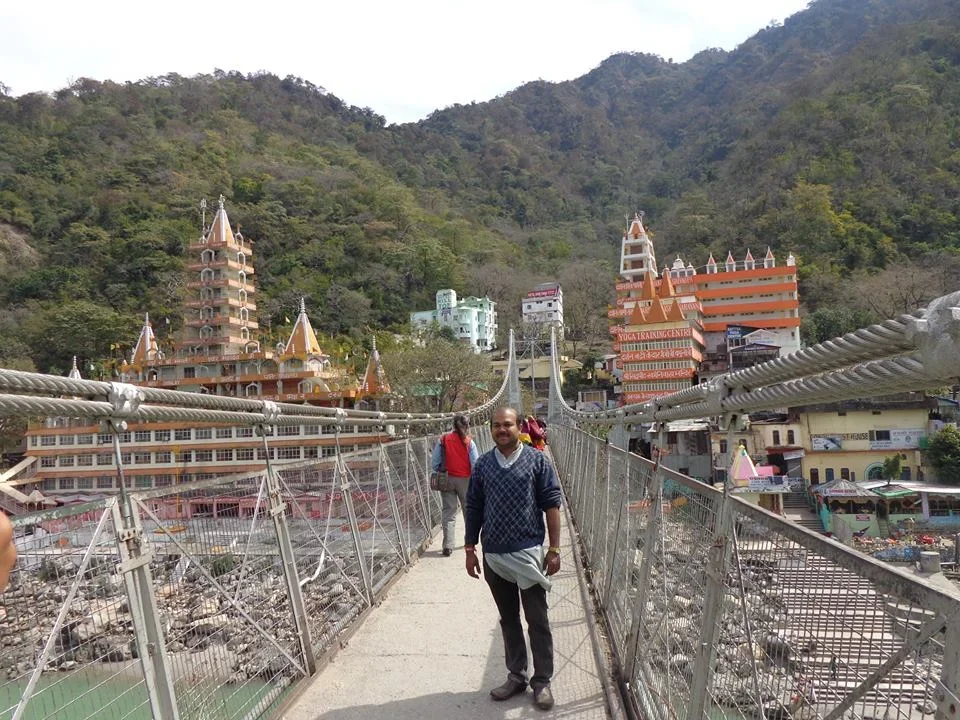 People walking across a suspension bridge with temples and buildings on a hillside in the background.