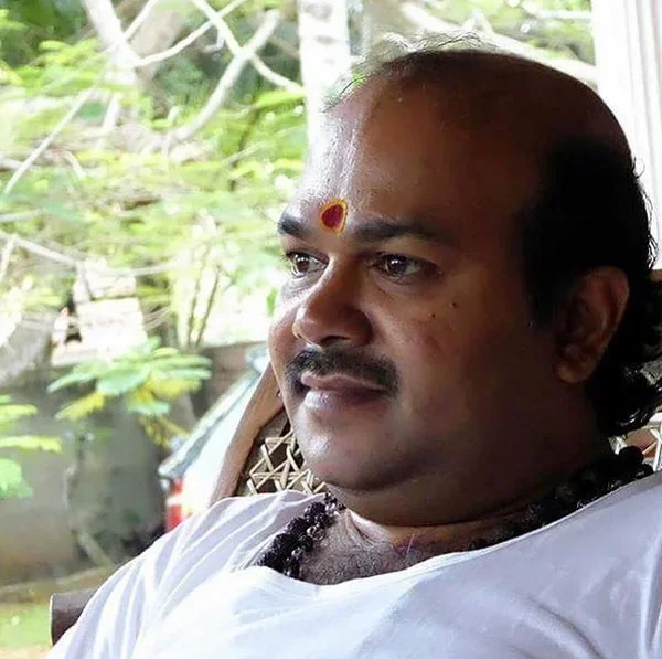 A man with a bindi on his forehead sitting in a room with greenery visible outside the window.