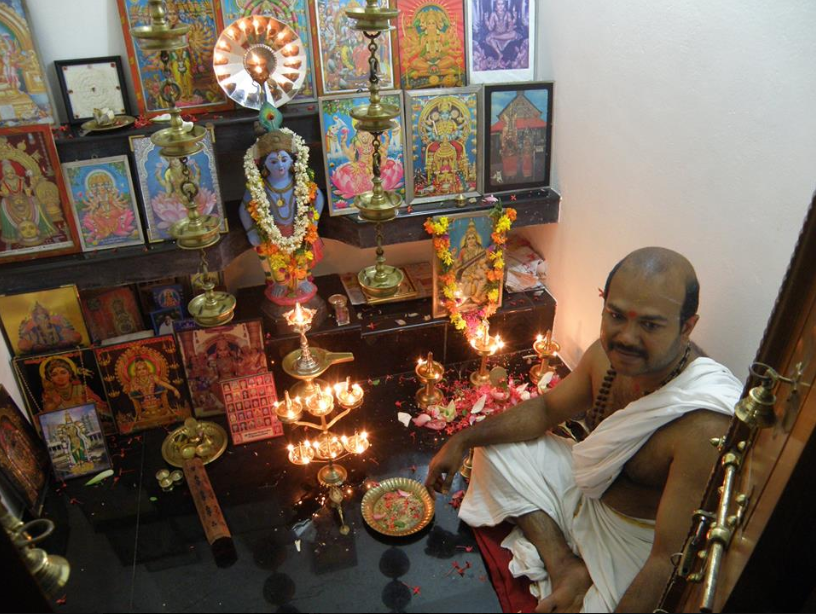 Hindu priest performing a ritual at a small shrine with framed images of deities, brass lamps, and offerings.