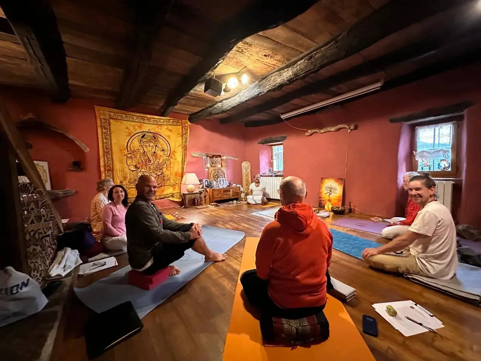 A group of people sitting on yoga mats and cushions in a cozy room decorated with spiritual art and artifacts, participating in a meditation or spiritual gathering.
