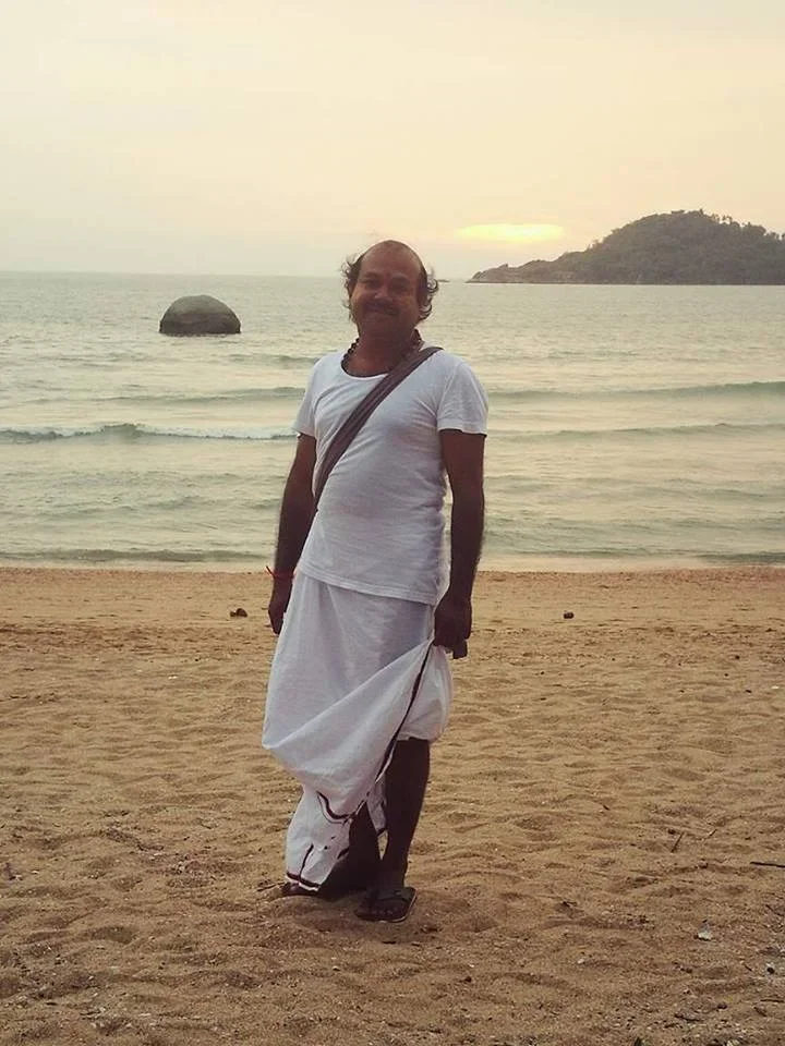 A man standing on a beach during sunset, wearing traditional white Indian attire with a bag slung across his shoulder, with an ocean and rocky island in the background.