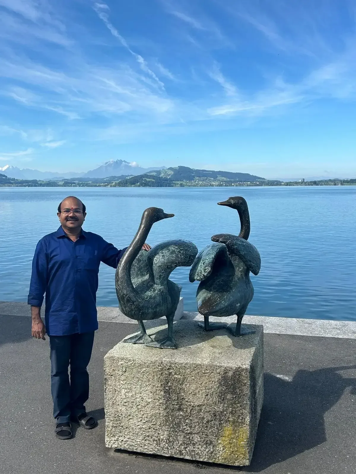 A man in a blue shirt and black pants standing next to two bronze sculptures of swans by a lake, with mountains and a blue sky in the background.