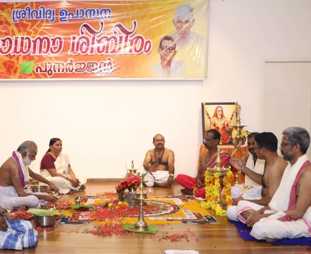 A traditional Hindu religious ceremony with multiple men and women sitting cross-legged on the floor, offering prayers to a central area with flowers, lamps, and religious items. There are portraits and religious icons in the background.