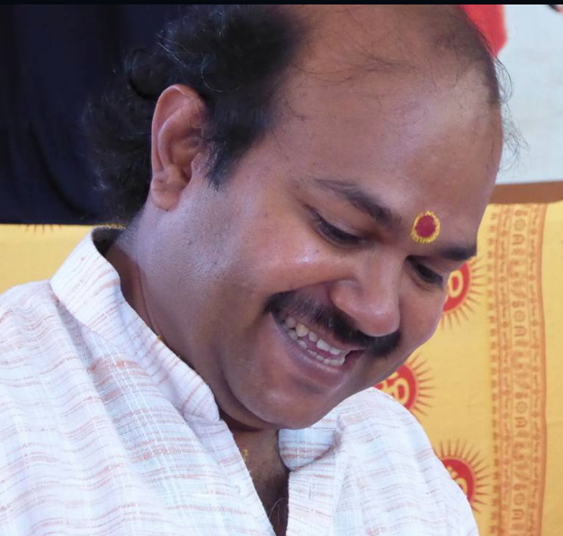 A smiling man with a mustache and a tilak on his forehead, wearing traditional Indian clothing, in front of yellow and red patterned background.