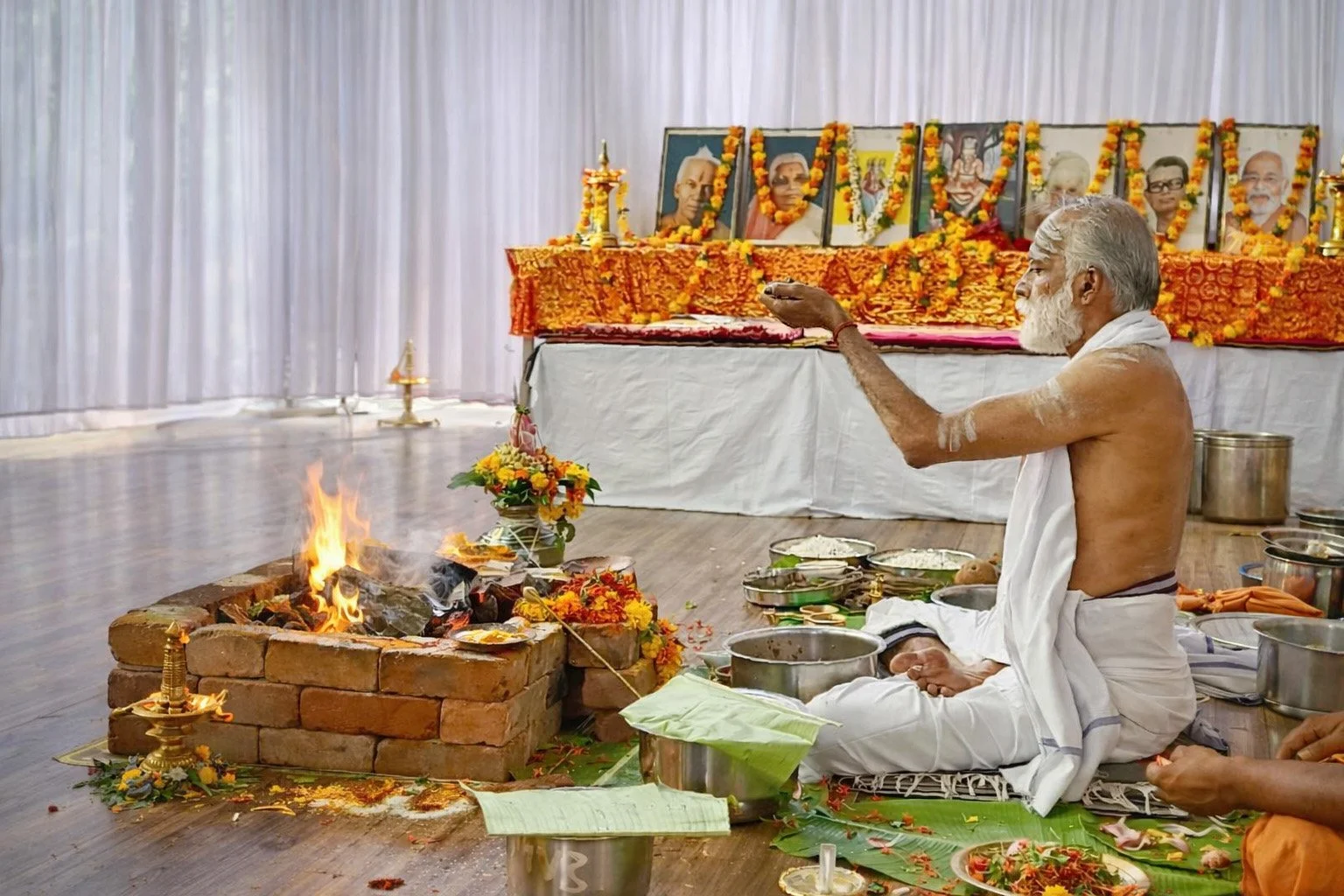 A Hindu priest performing a traditional fire ritual during a ceremony, with photographs of saints or gurus on a decorated altar in the background.