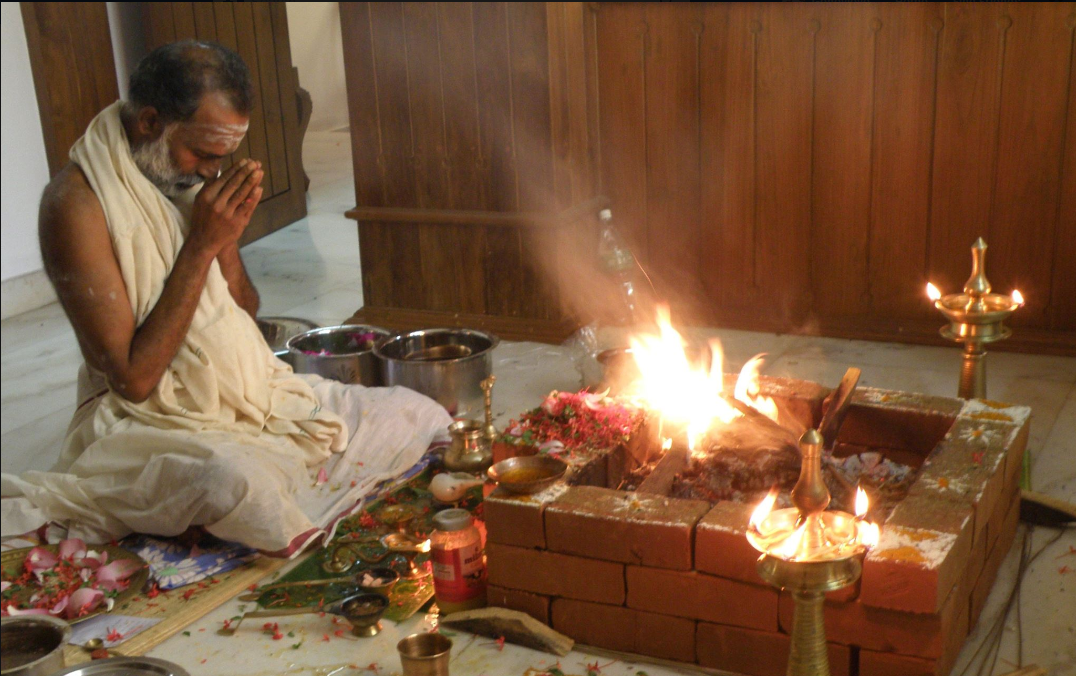 A man praying with folded hands in front of a fire in a traditional Hindu fire pit during a religious ceremony, surrounded by offerings and ritual items.