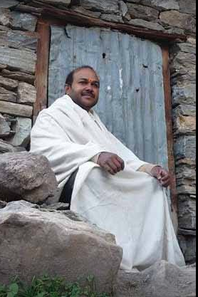 A man sitting on rocks outside a stone building with metal door, wearing a white shawl and smiling.