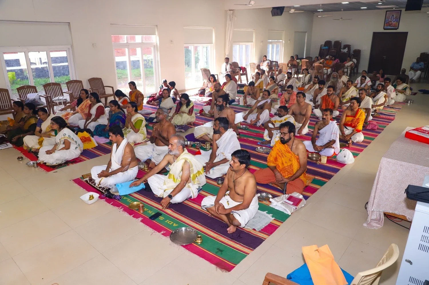 Hindu religious ceremony with men and women sitting on colorful rugs, wearing traditional attire, in a spacious hall with large windows, some men shirtless, participating in prayer or rituals.
