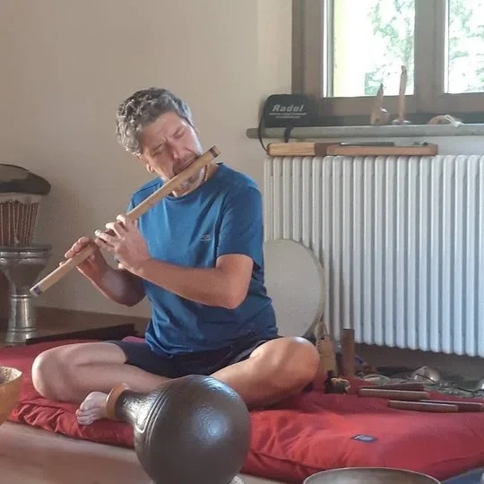 A boy sitting cross-legged on a red cushion indoors, playing a wooden flute with his eyes closed. There is a window behind him, a radiator, and various musical instruments and objects nearby.