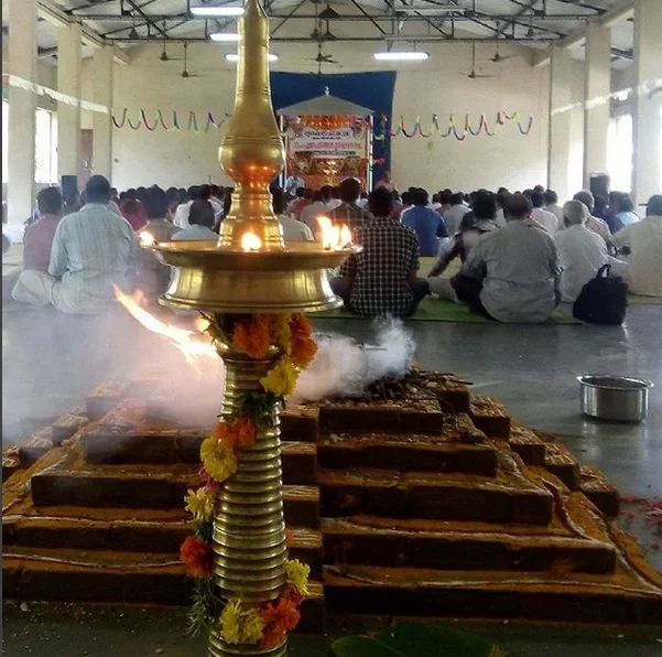 A large brass oil lamp decorated with flowers in the foreground, with a religious gathering of people sitting cross-legged on the floor in the background, inside a hall decorated with hanging streamers.