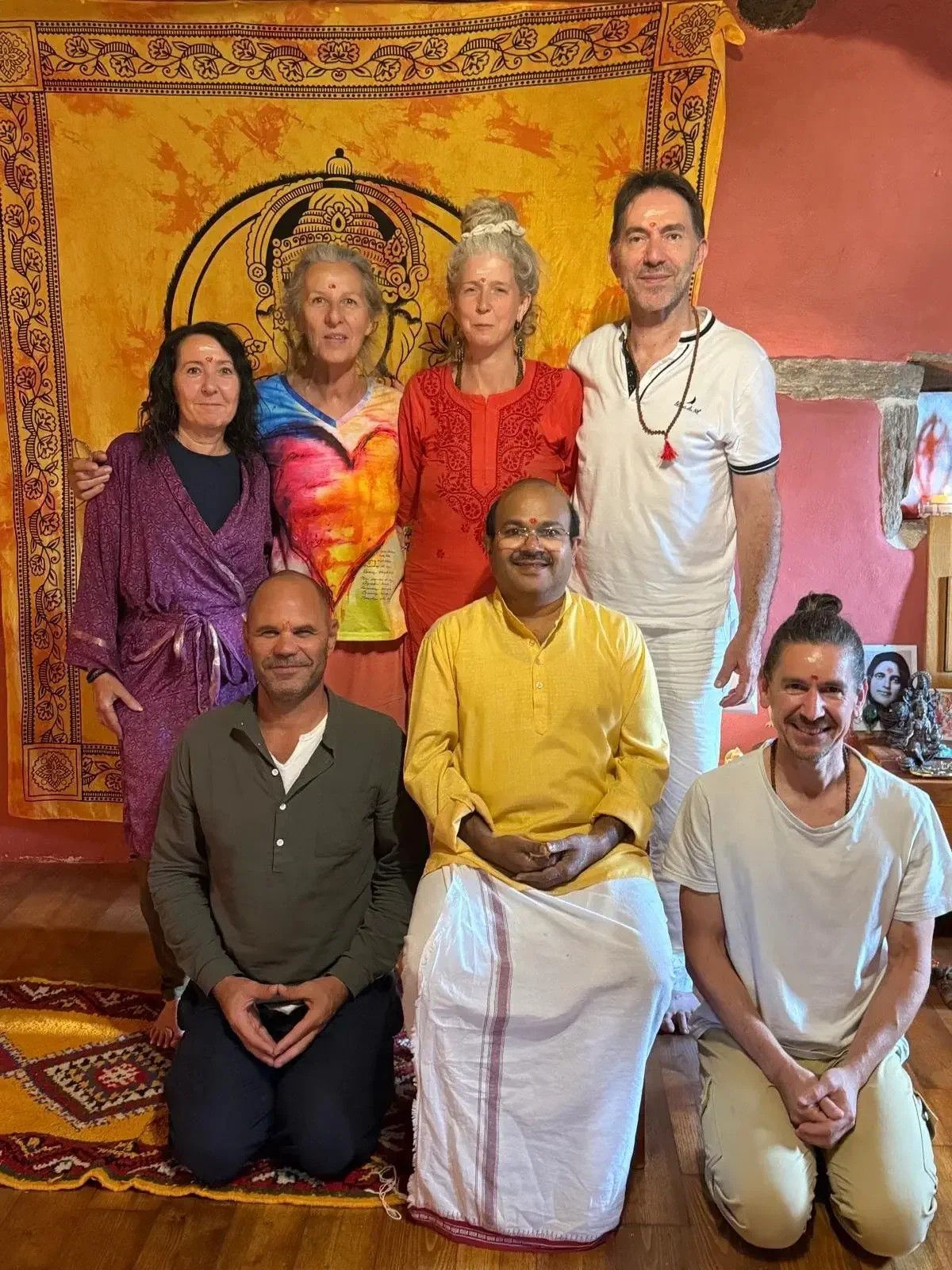 A group of eight people posing together indoors, with a yellow and orange tapestry depicting a deity or spiritual symbol behind them. They are dressed in colorful traditional and casual clothing, and some are sitting while others stand.