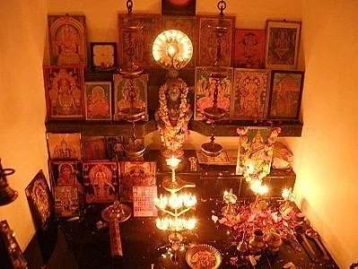 A small altar with multiple religious icons and statues, lit by a hanging light fixture, decorated with flowers and offerings.