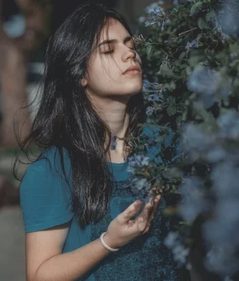 A young woman with long black hair and closed eyes standing among trees and foliage.