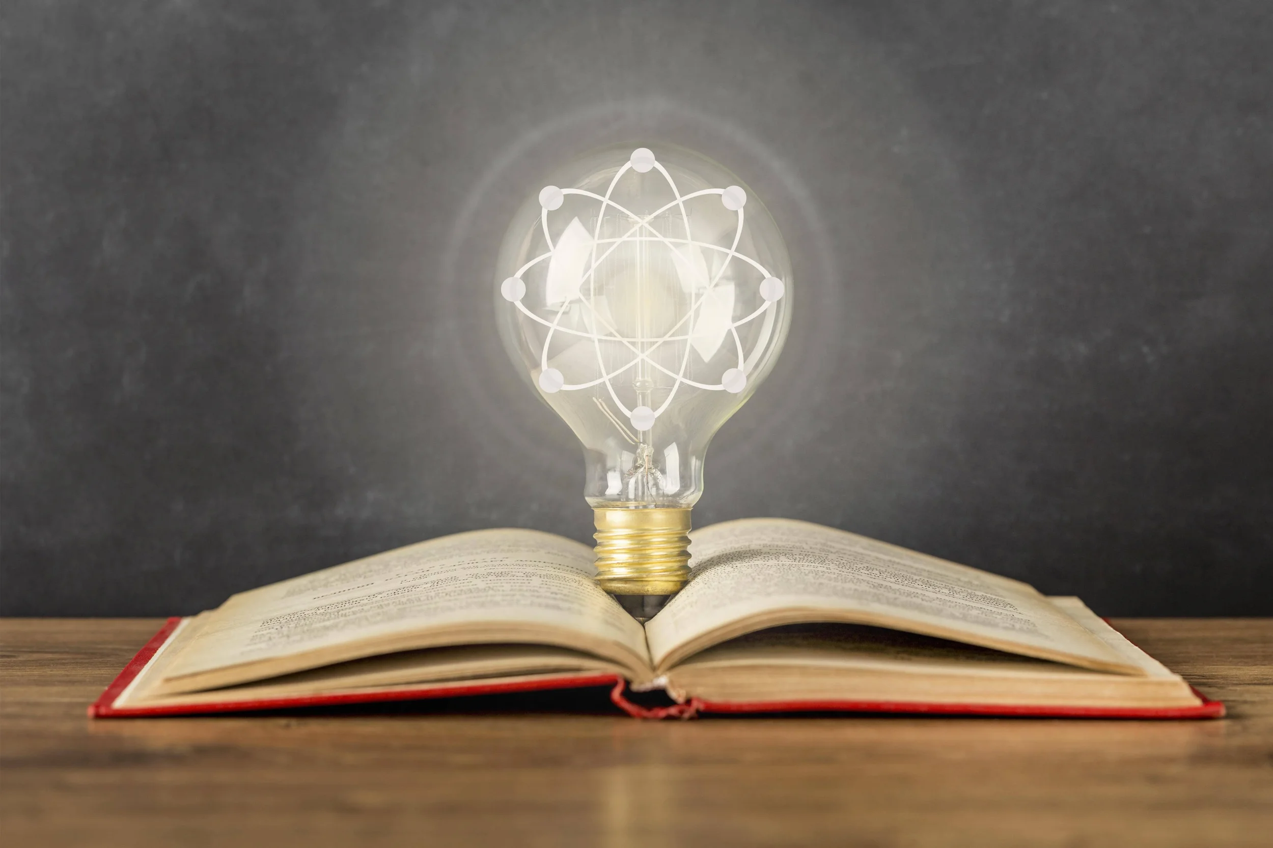 Open book with a lightbulb on top, the lightbulb has a graphic of an atom with electrons orbiting a nucleus on it.