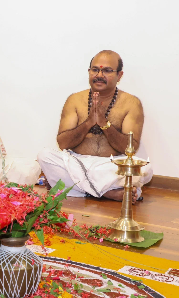 A man with glasses, a red mark on his forehead, and wearing traditional Indian attire sits cross-legged on the floor, with his hands folded in prayer. He is surrounded by flowers and a brass lamp, indicating a spiritual or religious ceremony.