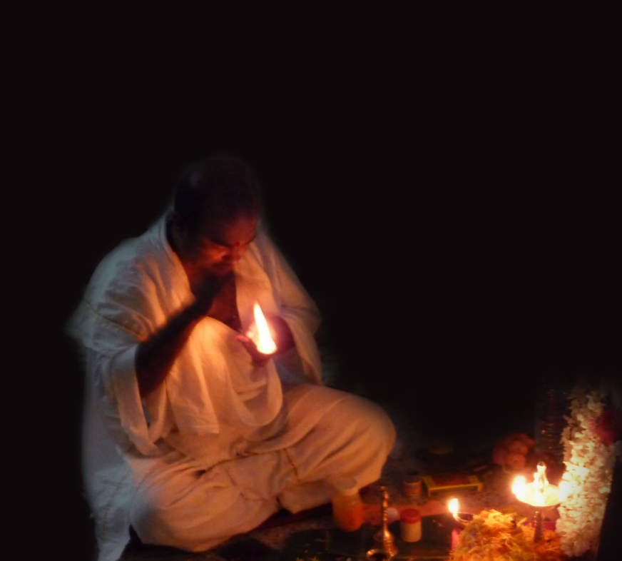An elderly man in traditional white attire, sitting cross-legged on the floor, performs a ritual with a flame in his hand during a religious ceremony in a dark environment.