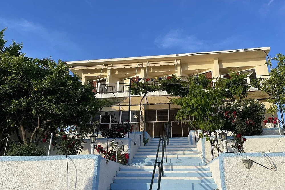 A white multi-story building with a staircase leading up to the entrance, surrounded by green trees and flowering plants, under a clear blue sky.
