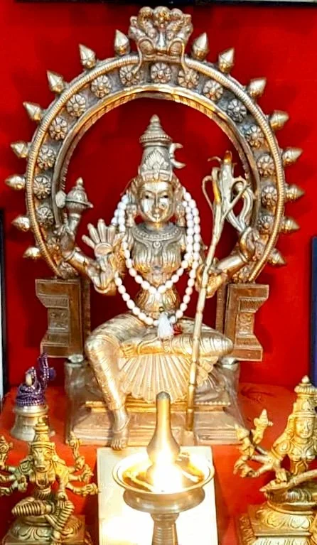 Hindu deity idol with a circular halo, seated on a throne, holding a staff, adorned with jewelry, surrounded by smaller statues, with a lamp in front, set against a red background.