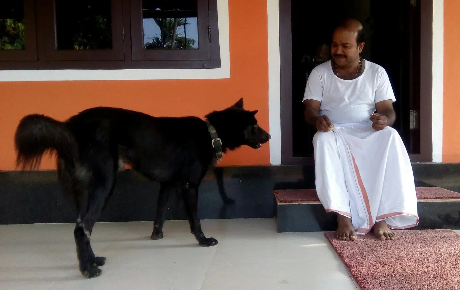 A man dressed in traditional white clothing sitting barefoot on a step, facing a black dog with a harness, outside a colorful house with orange walls and a dark window.