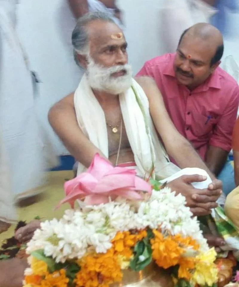 An elderly man with a white beard, traditional clothing, and a sacred mark on his forehead, seated with a bouquet of flowers in front of him. A man in a pink shirt is sitting close and conversing with him.