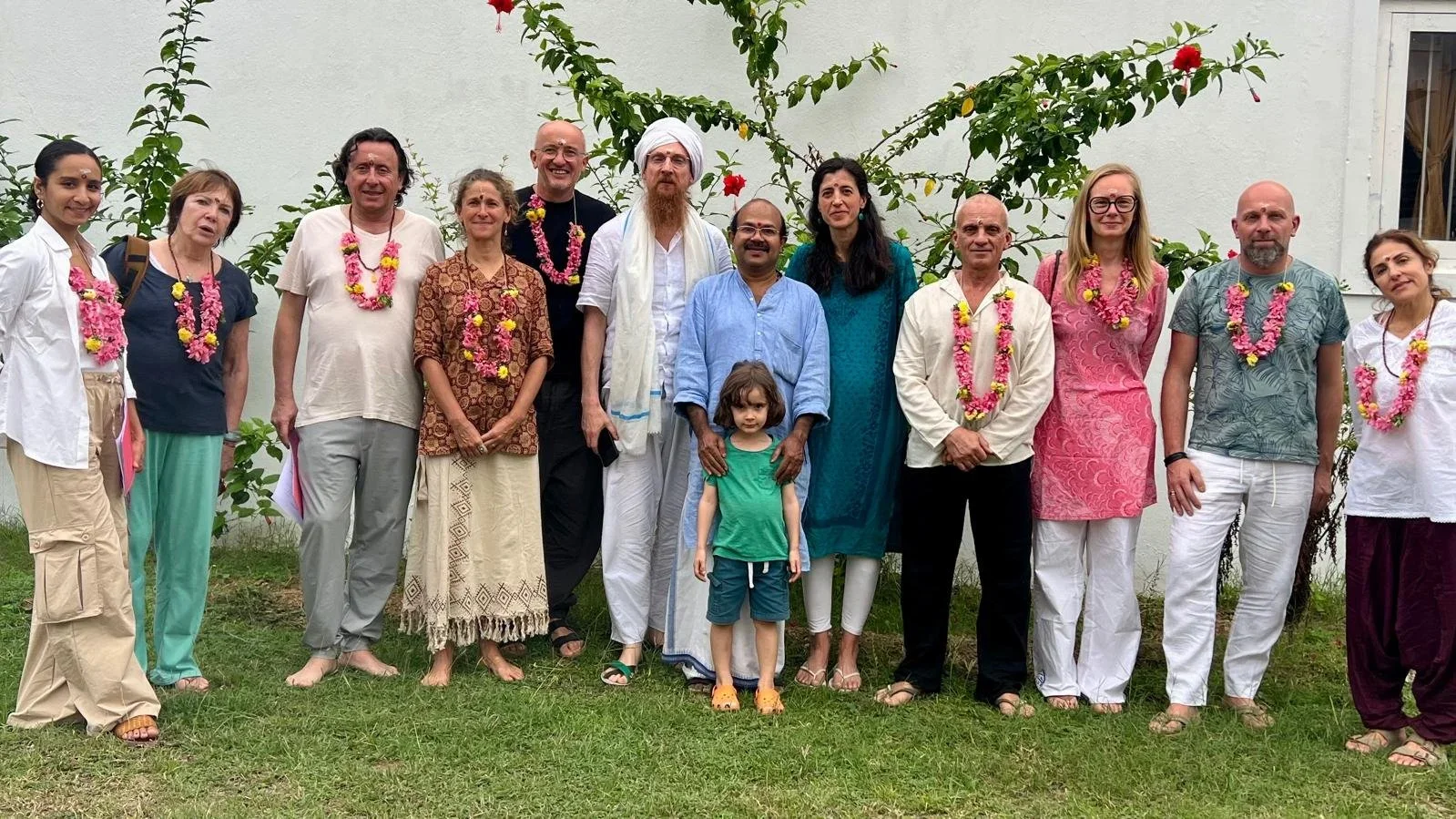 Group of people standing outdoors in front of a white wall and green plants, wearing traditional Indian attire and garlands of flowers, some smiling, some serious, including a young girl in a green shirt and a small child among adults.