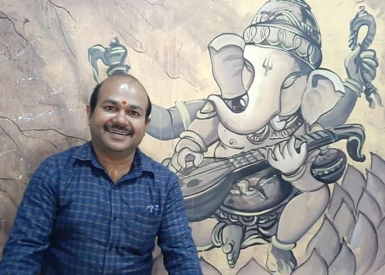 Man smiling in front of wall mural of Lord Ganesha playing a musical instrument.