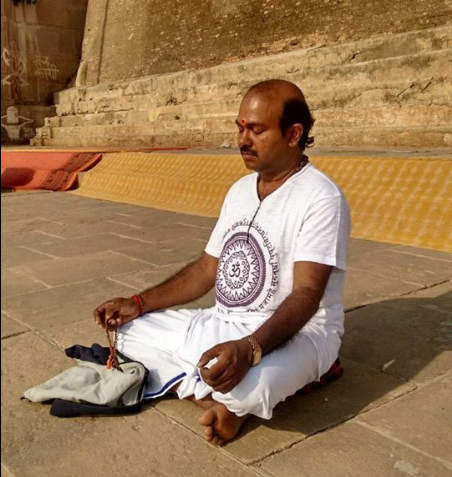 Man sitting cross-legged on stone steps praying or meditating with eyes closed, holding prayer beads in one hand, wearing white traditional clothing, outside near large steps.
