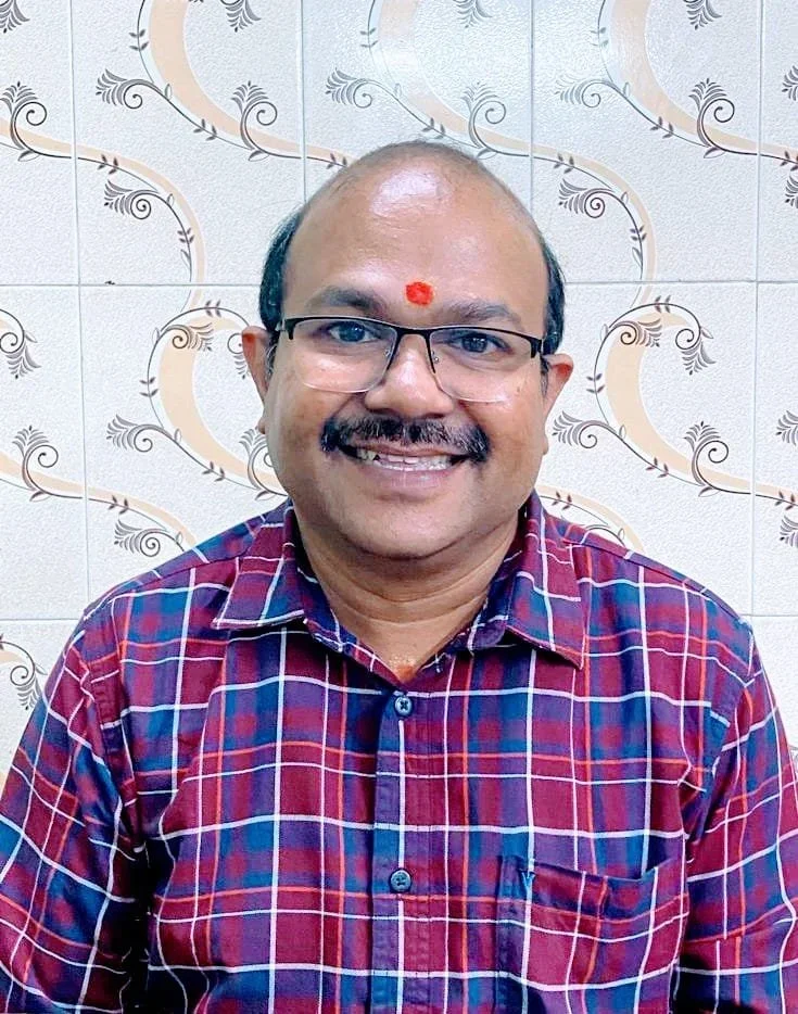 Man wearing glasses and a red, blue, and white checkered shirt, smiling, with a red mark on his forehead, against a tiled wall with decorative patterns.