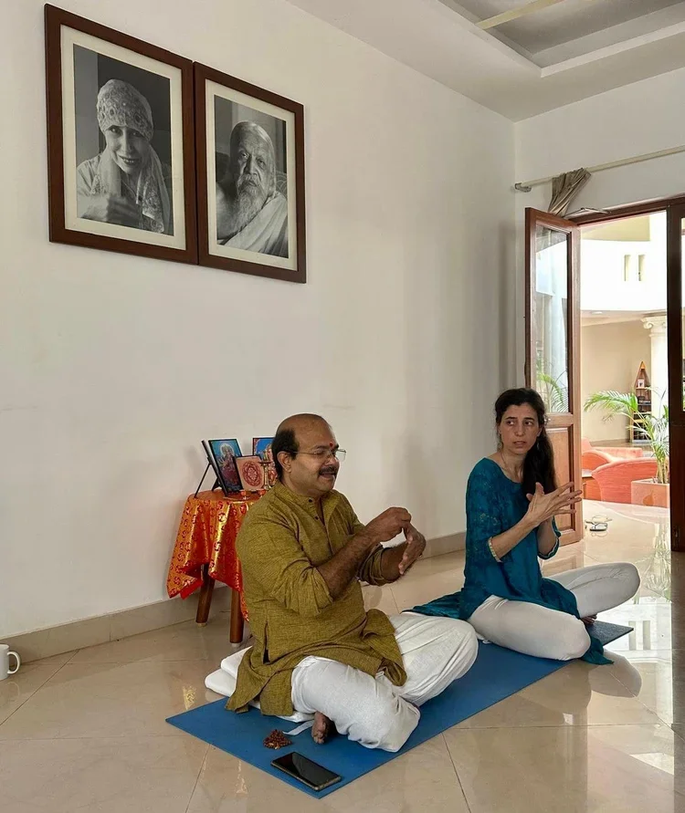 Two people sitting cross-legged on yoga mats in a room, engaged in a conversation or prayer. The man is smiling and holding prayer beads, while the woman is gesturing with her hands. There are framed black-and-white portraits on the wall, and a small