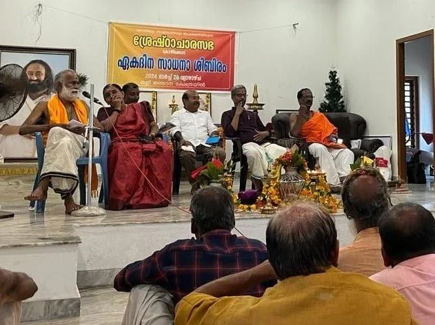 Panel of six men sitting on chairs at an indoor event, with people seated in the audience. A large portrait of a spiritual leader hangs on the wall to the left, and a colorful banner with text in a South Indian language is behind the panel. The floor