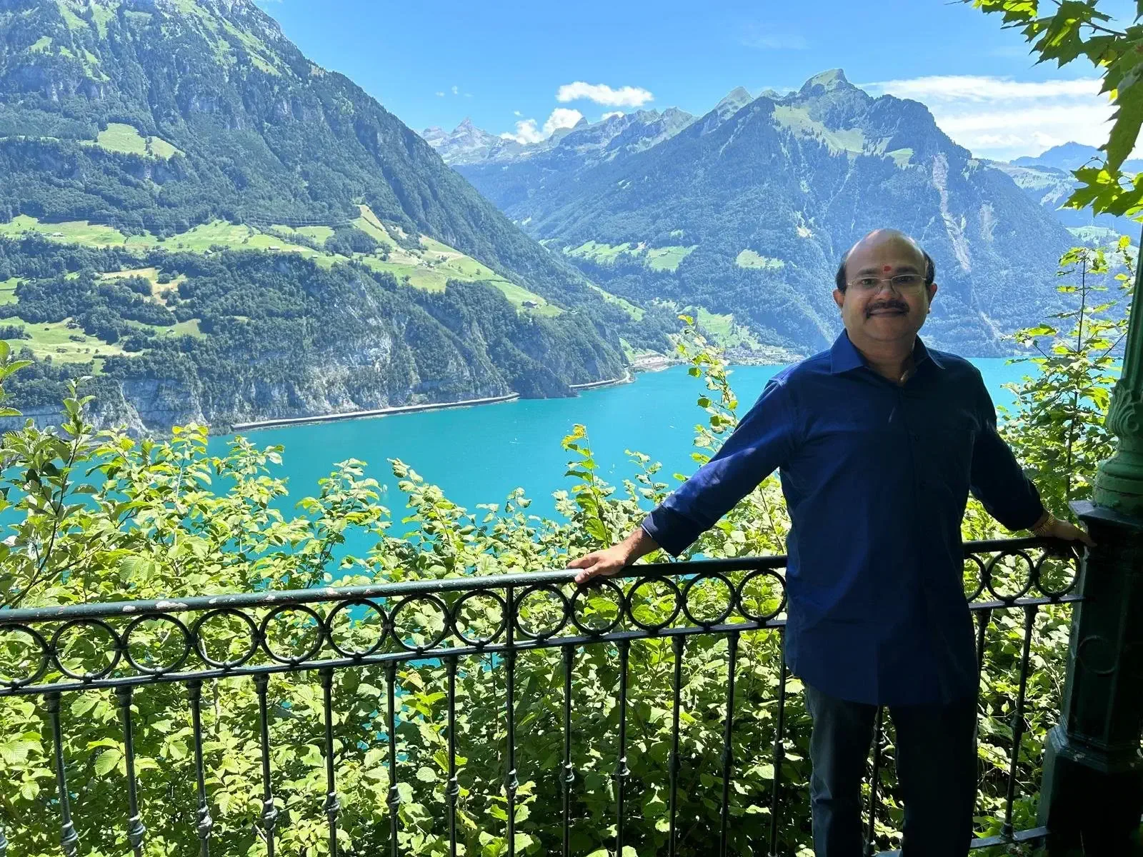 A man with glasses and a mustache wearing a blue shirt standing on a balcony with a scenic view of a large lake, mountains, and green hills and trees in the background.