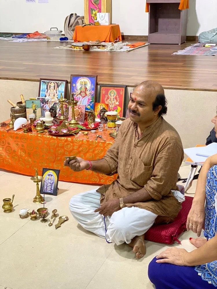 A man sitting cross-legged on the floor in front of a decorated altar with religious images, brass lamps, and offerings, during a Hindu prayer or ritual.