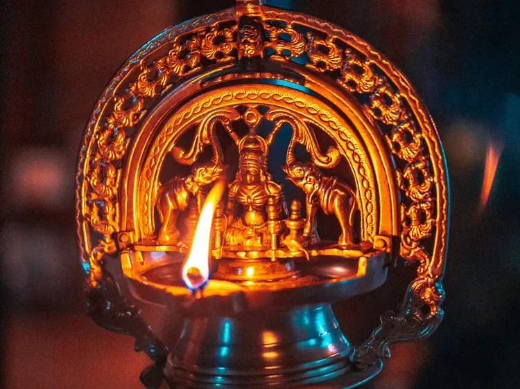 A brass or bronze lit oil lamp with intricate carvings and a central deity figure, surrounded by decorative elements, glowing with a small flame in a dark setting.