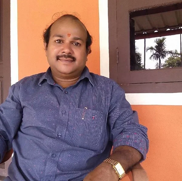 A man with a mustache and balding head sitting indoors, wearing a blue shirt, gold watch, and a red dot on his forehead, with an orange wall and window showing palm trees in the background.