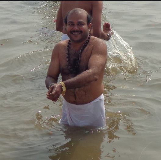 Man smiling, wearing a white garment, standing in water, with another person partially visible behind him.