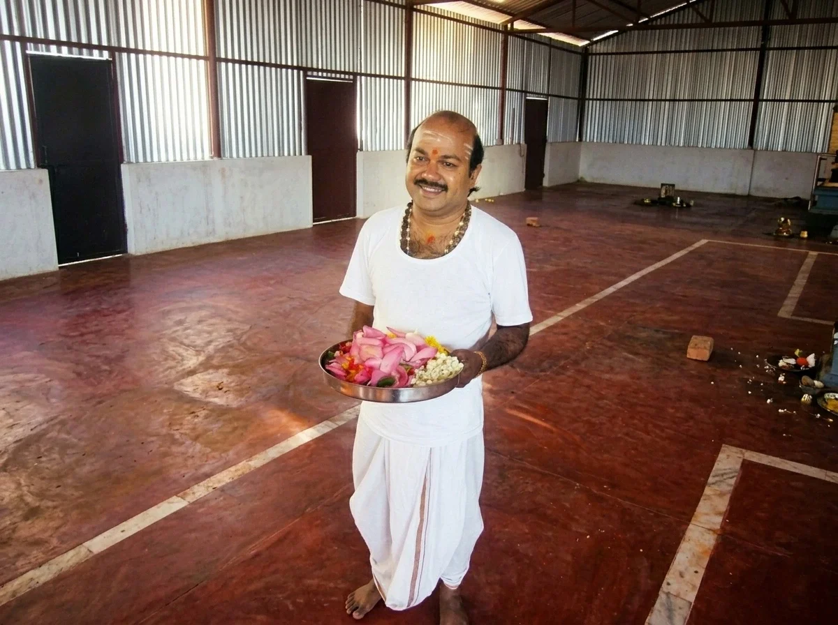 A man standing barefoot in a large, empty hall with metal walls, holding a metal tray with flower petals, flowers, and turmeric roots, smiling at the camera.