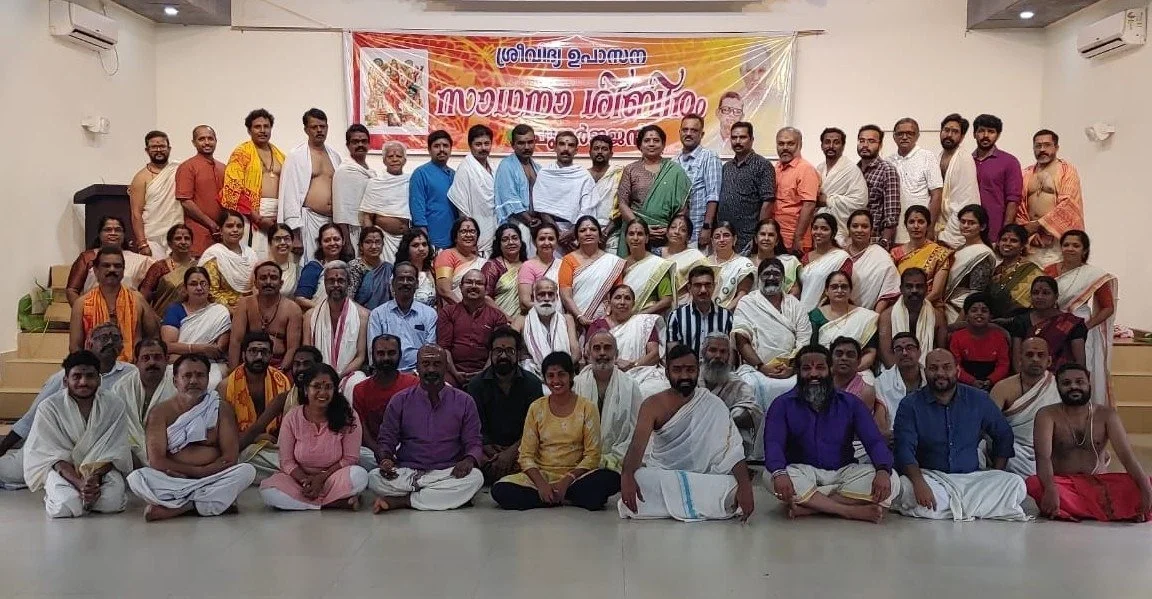 Group photo of people, including men and women, some in traditional Indian attire, gathered in a hall for an event or celebration. There is a banner with Malayalam text in the background.