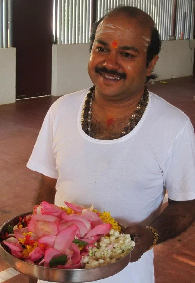 A man with a mustache, wearing a white shirt, holding a tray with flower petals and garlands, in a room with metallic walls and wooden floor.