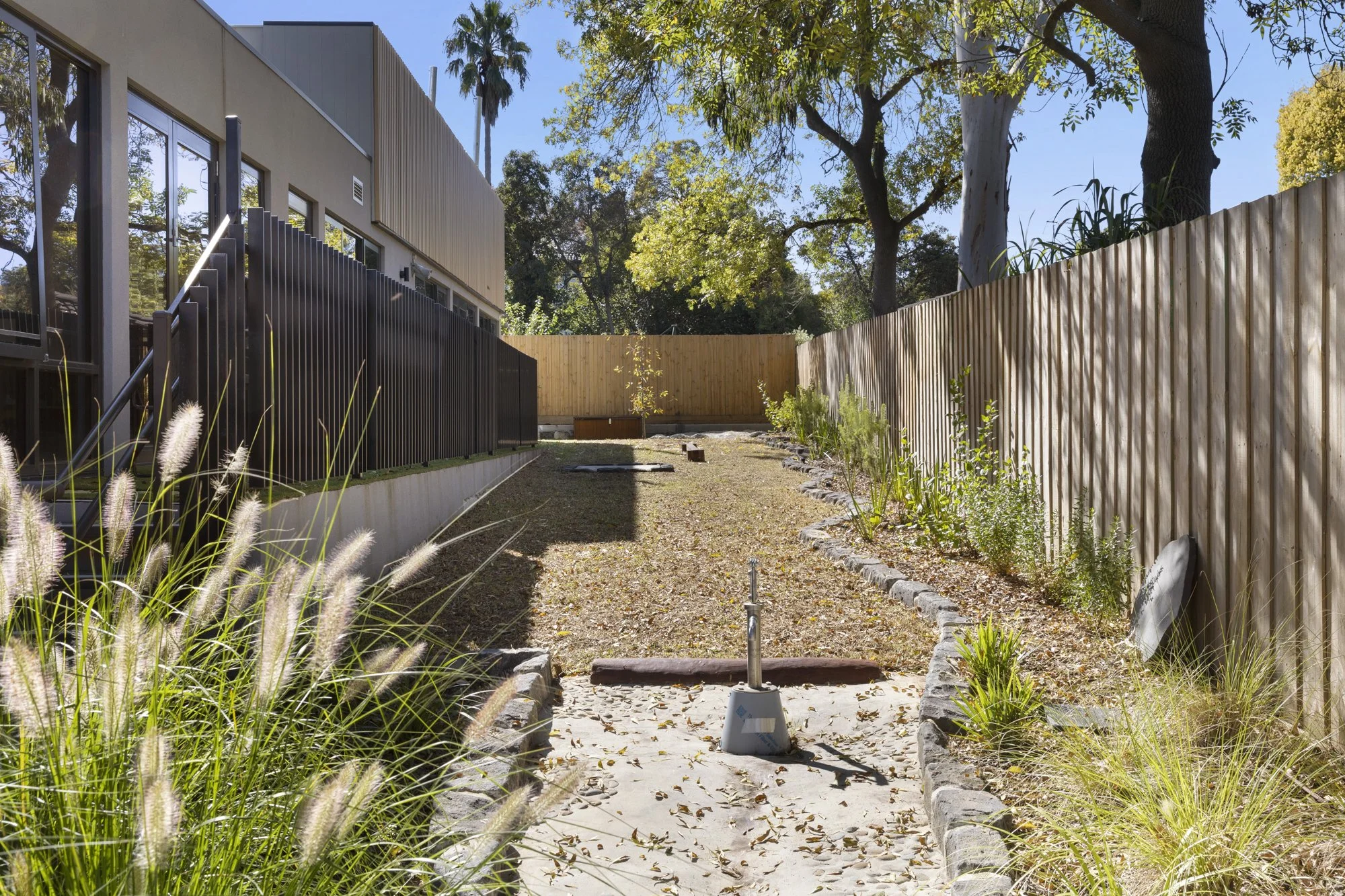 A backyard with a concrete path, grass and plants on the sides, a wooden fence, and trees with green leaves. There is a building with large windows and a black fence on the left side.
