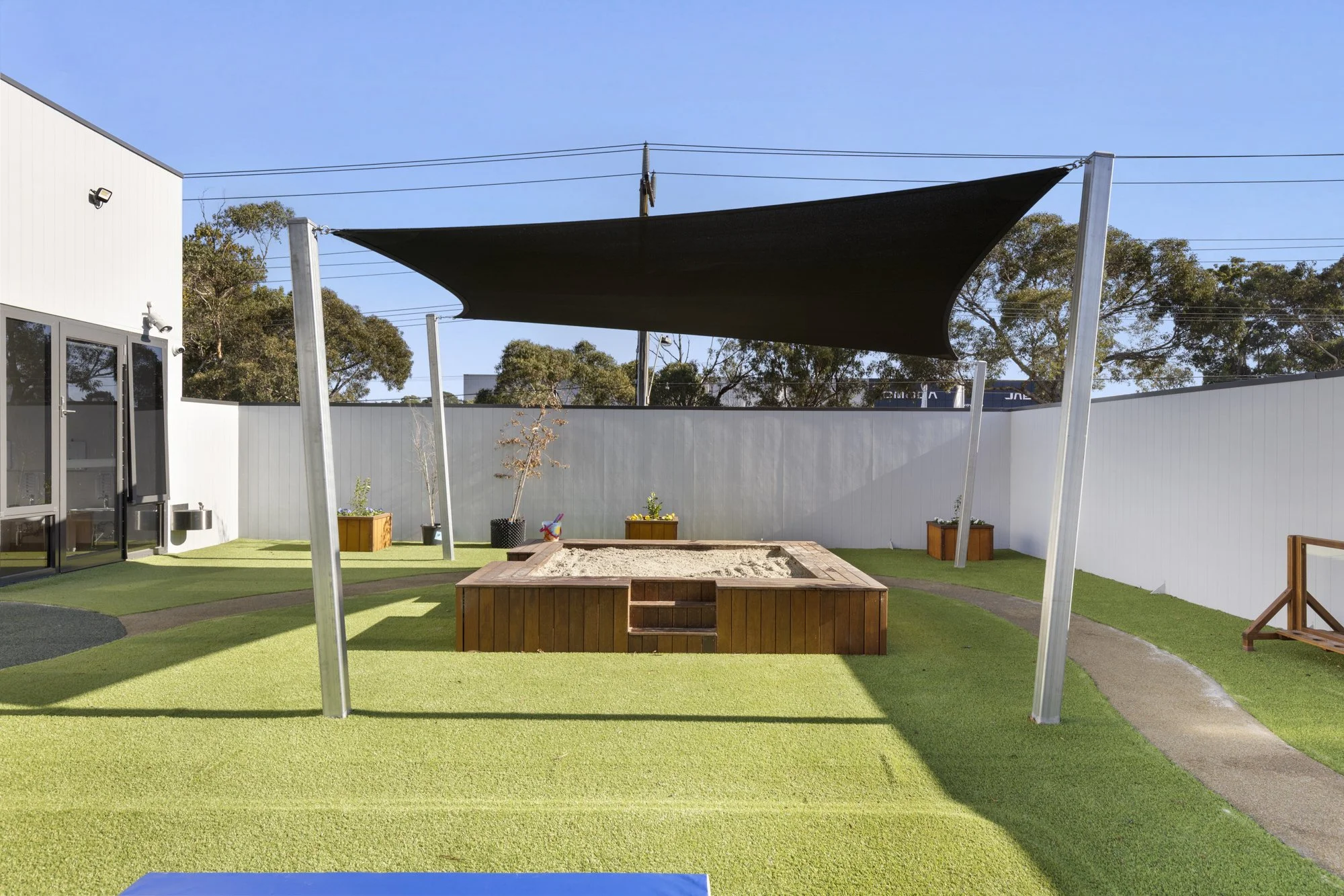 Outdoor play area with a sandbox, small trees in planters, shaded by a large black canopy, fenced with white walls, with a building on the left and trees in the background.